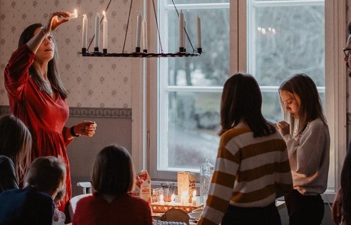 Barn runt ett bord med julmat. Amanda tänder ett ljus i ljuskronan ovanför bordet