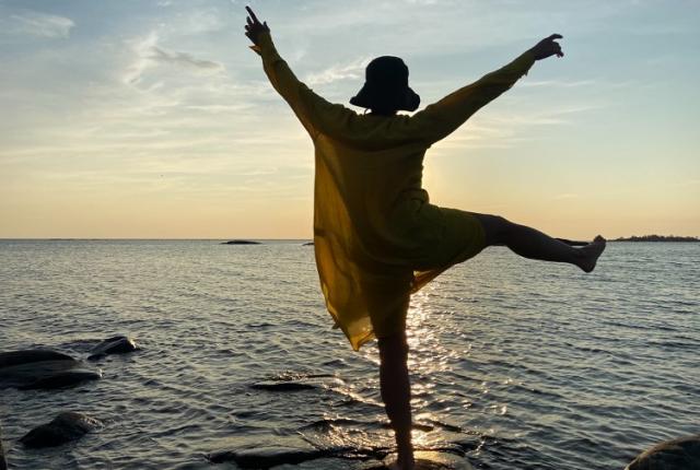 En kvinna vid havet i solnedgång. Hon blickar ut över horisonten och har händerna och ena benet lyft i glädje.