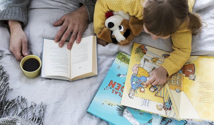 Ett barn ligger på mage och läser bilderböcker. Bredvid ser man händerna av en vuxen med en egen bok och en kaffekopp.