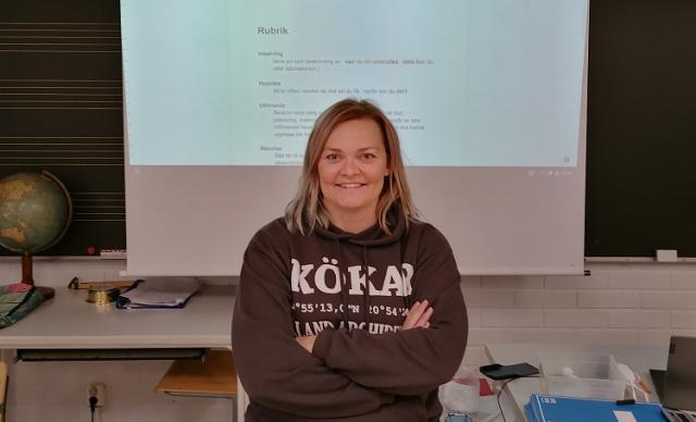 Lärare Cecilia Österlund i klassrummet. I bakgrunden ser man en presentation av en labbrapport.