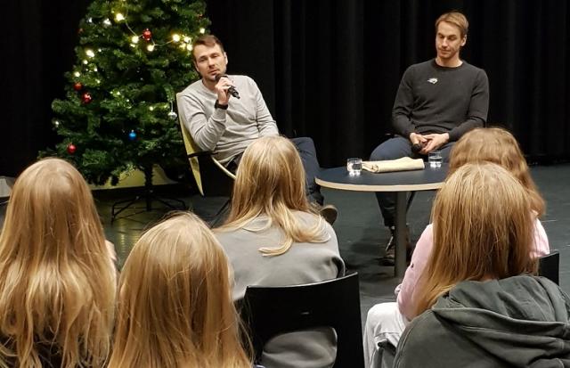 Textförfattaren Mikael Gros sitter vid ett bord och intervjuar ishockeyspelaren Erik Riska. I förgrunden ser man huvuden av publiken, elever i åk 5. I bakgrunden ses en julgran.