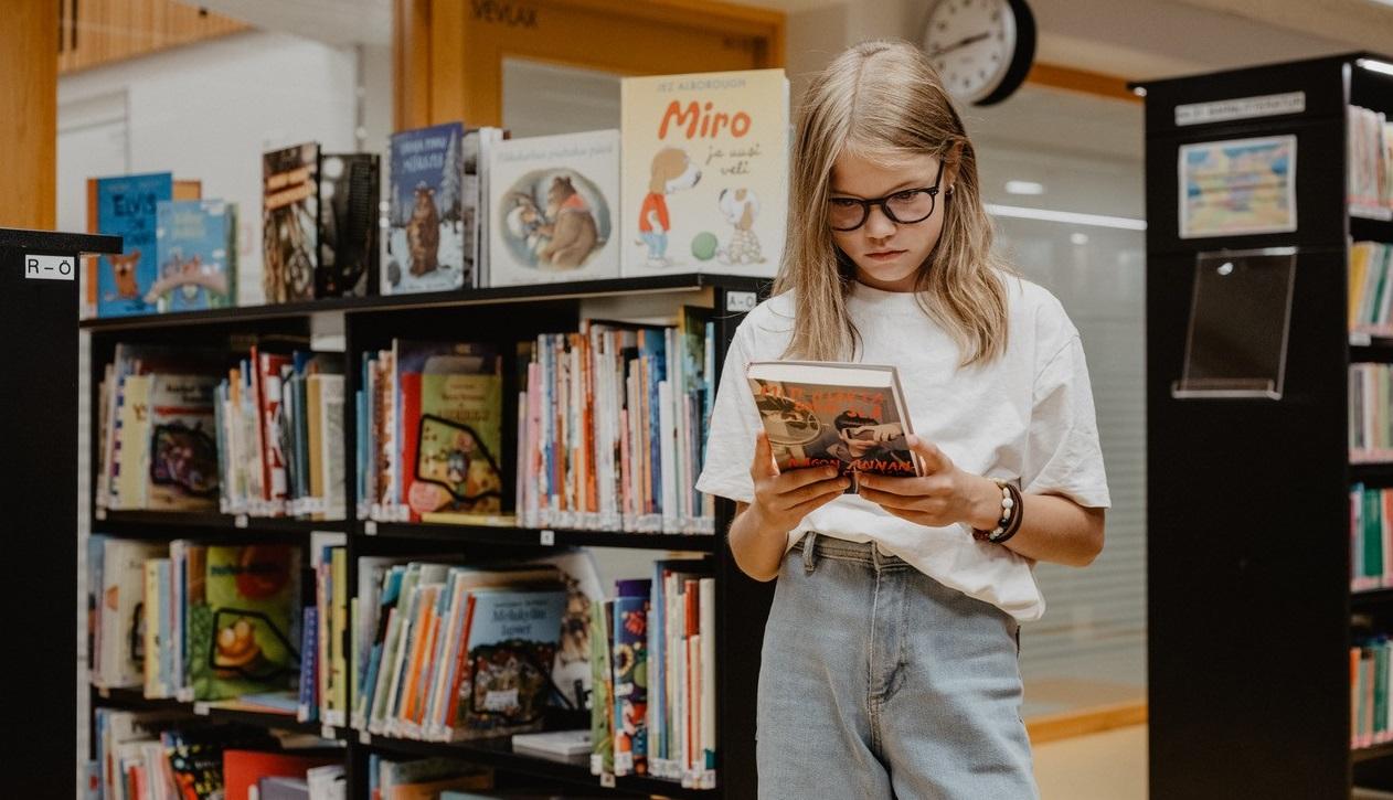 En flicka står i ett bibliotek med en bok i handen.